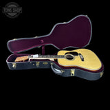 An acoustic guitar with a natural wood top and dark neck, displayed inside an open black hard case with a deep purple velvet interior, set against a solid black background.