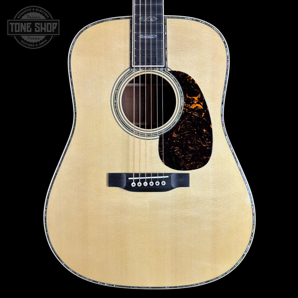 A front-facing acoustic guitar with a natural wood finish and a dark tortoiseshell pickguard, shown against a solid black background with a Tone Shop watermark in the top-left corner.