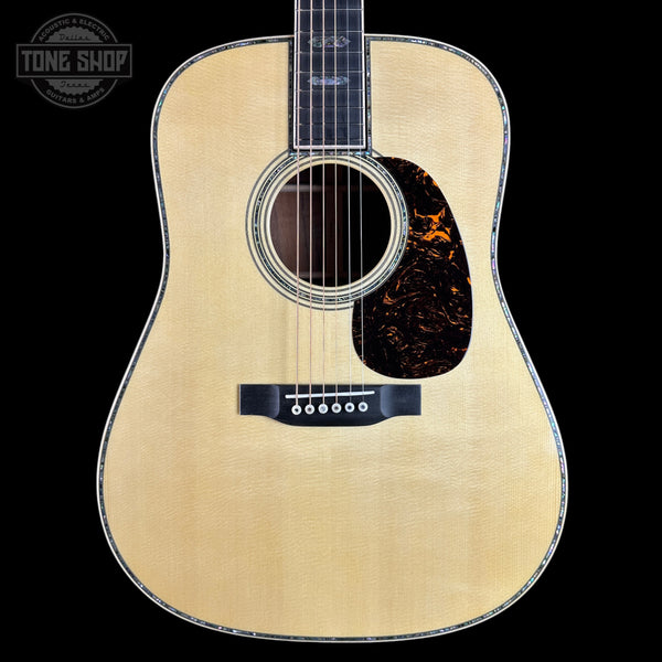 A front-facing acoustic guitar with a natural wood finish and a dark tortoiseshell pickguard, shown against a solid black background with a Tone Shop watermark in the top-left corner.