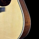 Close-up detail of an acoustic guitar body showing the natural wood top, a dark brown wood side, and a black pickguard against a solid black background.