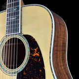 Close-up of an acoustic guitar body showing the natural wood soundboard, dark brown wood side, and a black tortoiseshell-style pickguard against a black background.
