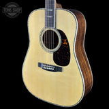 An acoustic guitar with a natural wood top and a dark brown wood side, shown against a solid black background. A circular 'TONE SHOP' watermark is visible in the upper left corner.
