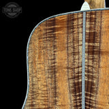Close-up of the back of an acoustic guitar showing a rich figured wood grain finish in warm brown and amber tones, with a decorative metal downpin and a black background. A Tone Shop watermark is visible in the top-left corner.