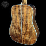 Close-up of the back of an acoustic guitar showing a rich, figured wood grain pattern in warm brown and golden tones, with a silver-colored metal edge trim, set against a solid black background.