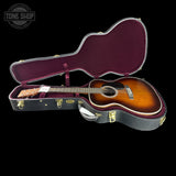 An acoustic guitar with a sunburst finish resting inside an open black hard case with a burgundy interior, shown against a solid black background. A Tone Shop watermark is visible in the top-left corner.
