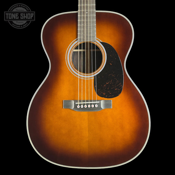 A close-up studio shot of an acoustic guitar body against a black background, showing a natural sunburst finish that fades from bright amber-orange at the edges to a darker brown center, with a black pickguard and white binding.