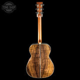 The back of a wooden acoustic guitar with a prominent figured wood grain pattern, shown against a solid black background with a Tone Shop watermark in the top-left corner.