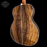 The back of an acoustic guitar made from a figured wood species with dramatic wavy grain patterns, featuring a dark brown and golden-brown wood finish with white binding, shown against a solid black background.