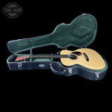 An acoustic guitar with a natural wood body and dark neck, resting inside an open black hard case with green felt lining, shown against a black background. A Tone Shop watermark is visible in the top left corner.