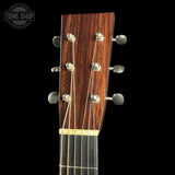 Close-up of the headstock of an acoustic guitar with a dark brown wood grain finish, six silver tuning pegs, and six string tuners, shown against a solid black background with a Tone Shop watermark in the top left corner.