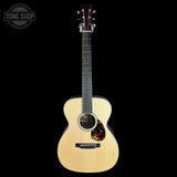 A natural wood acoustic guitar with a dark headstock, displayed vertically against a solid black background. A circular 'TONE SHOP' watermark is visible in the top-left corner.