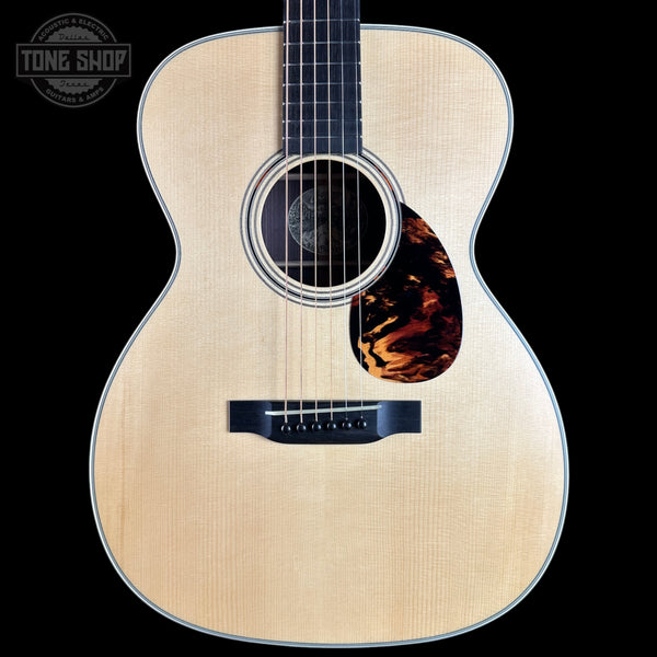 An acoustic guitar with a natural wood finish and a tortoiseshell pickguard, shown against a solid black background with a Tone Shop logo in the top-left corner.