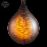Close-up of a teardrop-shaped wooden headstock or lamp-like wooden object with a glossy, flame-grained brown finish, glowing with warm amber light against a black background. A Tone Shop watermark is visible in the top-left corner.