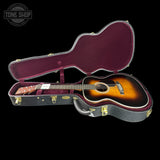 An acoustic guitar with a sunburst finish displayed inside an open hard case with a burgundy velvet interior, set against a solid black background.