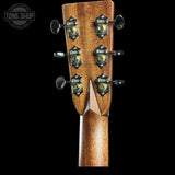 Close-up of the headstock of an acoustic guitar, showing six tuning pegs with decorative black and gold covers and a warm brown wood grain finish, set against a solid black background.
