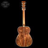 A wooden acoustic guitar shown from the back, featuring a rich, wavy wood grain pattern on the backplate and neck, set against a solid black background with a Tone Shop watermark in the top-left corner.