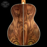 Close-up of the back of an acoustic guitar showing a rich, wavy wood grain pattern in warm brown and amber tones, set against a solid black background with a Tone Shop watermark in the top left.