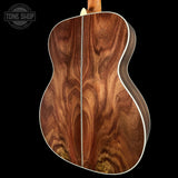 Close-up of the back of an acoustic guitar showing a rich, swirling natural wood grain pattern in warm brown and amber tones, set against a solid black background with a Tone Shop watermark in the top left.