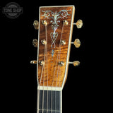 Close-up of an acoustic guitar headstock featuring a richly grained brown wood finish, ornate silver inlay, and gold tuning pegs, shown against a solid black background with a Tone Shop watermark in the top left.