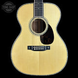 A close-up studio shot of an acoustic guitar body against a solid black background, featuring a natural wood finish, a dark pickguard, and a Tone Shop watermark in the upper left corner.
