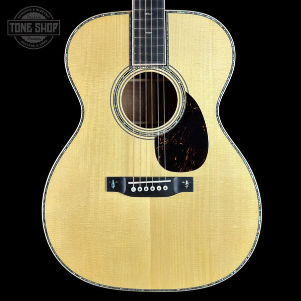 A close-up studio shot of an acoustic guitar body against a solid black background, featuring a natural wood finish, a dark pickguard, and a Tone Shop watermark in the upper left corner.