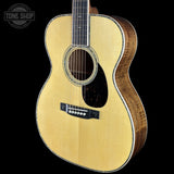 A close-up studio shot of an acoustic guitar body against a solid black background, showing a natural wood finish, sound hole, pickguard, and bridge. A Tone Shop watermark is visible in the top-left corner.