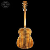 The back of an acoustic guitar with a highly figured warm brown wood finish, shown against a solid black background with a Tone Shop watermark in the top-left corner.