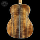 The back of an acoustic guitar made of figured wood, showing a rich brown and golden grain pattern, photographed against a solid black background with a Tone Shop watermark in the top left corner.