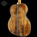 Close-up of the back of an acoustic guitar showing a rich, figured wood grain top in warm brown and amber tones, with white binding and a visible Tone Shop watermark in the upper left corner.