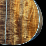 Close-up of the back of an acoustic guitar showing a rich, figured wood grain finish in warm brown and golden tones, with a silver metal edge and a white inlay line.