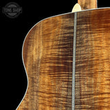 Close-up of the back of an acoustic guitar showing a richly figured flamed wood top in warm brown and amber tones, with a decorative inlay running vertically and a white edge binding against a black background.