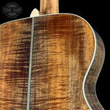 Close-up of the back of an acoustic guitar showing a rich, figured maple wood finish with a silver-tone soundhole rosette and white binding, set against a solid black background.