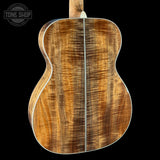 The back of a wooden acoustic guitar, showcasing a rich figured top with warm brown and golden tones, a central sound hole, and white binding, set against a solid black background with a Tone Shop logo in the top left corner.