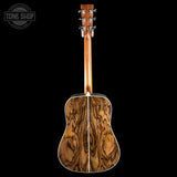 The back of an acoustic guitar with a rich, wavy wood grain pattern on the soundboard, shown against a solid black background. A Tone Shop watermark is visible in the top-left corner.