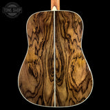 Close-up of the back of an acoustic guitar showing a rich, symmetrical wood grain pattern in warm brown and golden tones, set against a solid black background with a Tone Shop watermark in the top left corner.