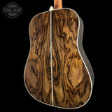 Close-up of the back of an acoustic guitar showing a rich, wavy wood grain pattern in shades of brown and golden tan, with a white binding edge against a solid black background.