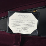 A black wooden presentation case containing a white Certificate of Authenticity for a Martin Custom Shop guitar, placed on a dark burgundy velvet surface.