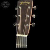 Close-up of the headstock of a Martin & Co acoustic guitar against a black background, showing the dark wood finish, gold script logo, six tuning pegs, and part of the neck with six strings.