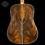 Close-up of the back of an acoustic guitar showing a rich, symmetrical wood grain pattern in warm brown and golden tones, with a white binding edge against a black background.