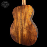The back of an acoustic guitar with a rich, warm brown wood grain finish, shown against a solid black background with a Tone Shop watermark in the top left corner.