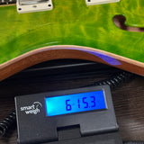 A close-up of a green electric guitar body with a digital smart weigh scale placed underneath the edge to measure the wood tone and curvature. The scale displays a weight reading of 6.
