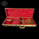 A gold-finish electric guitar resting inside an open hard case with bright red plush lining, shown against a solid black background. A Tone Shop watermark is visible in the top-left corner.