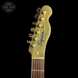 Close-up of a gold-finished electric guitar headstock with gold tuning pegs, black script branding, and a dark wood fretboard visible against a black background.