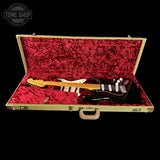 A black electric guitar with a yellow maple neck and white headstock is displayed inside an open vintage-style hard case with a red plush lining, set against a solid black background.
