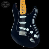 A black electric guitar with a maple neck, featuring a glossy black finish, chrome hardware, and a white pickguard, shown against a black background with a Tone Shop watermark.