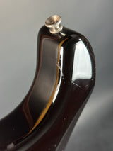 Close-up of the glossy dark brown headstock of an electric guitar, showing a metallic gold binding strip and a silver tuning peg against a gray background.