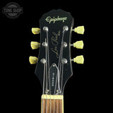 Close-up of the headstock of an Epiphone Les Paul Custom electric guitar against a black background, showing the brand name, model, six tuning pegs, and part of the fretboard.