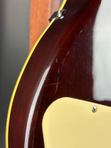 Close-up of the lower bout corner of an electric guitar body, showing a glossy dark reddish-brown finish, a cream pickguard, and a thin yellow binding edge.