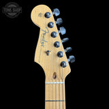 Close-up of a Fender Stratocaster electric guitar headstock in a natural wood finish, featuring six black tuning pegs and the Fender logo, set against a solid black background.