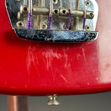 Close-up of a red electric guitar body near the bridge, showing a silver metal bridge assembly with purple string winds and visible surface scratches on the glossy red finish.